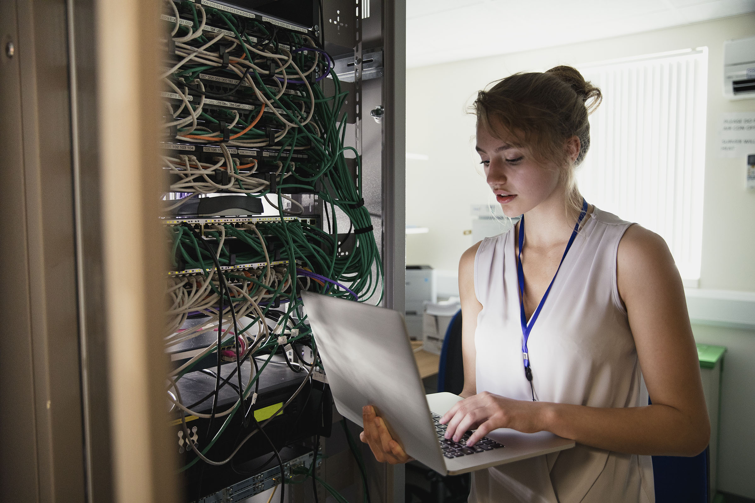 woman standing in an office on the right of image holding a laptop typing with one hand near a shelf full of servers with hundreds of cables connected to each other showing the importance of cyber security services Data Center Cybersecurity