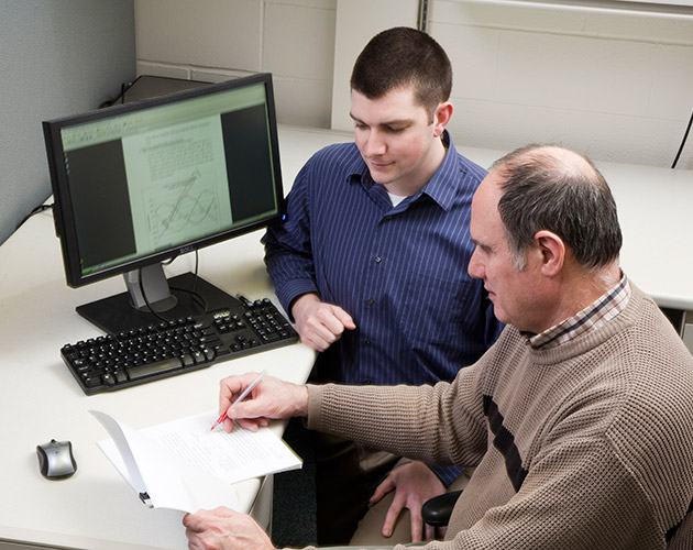 an older man planning on paper and a younger man watching him write while both sitting in front of a desk with a computer showing a synode team members working together, working infront of a computer, viewing a synode on computer, synode