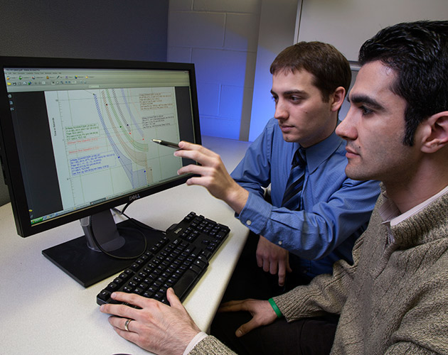 a man pointing at a computer screen with a pen wearing a blue shirt and dark tie to a man looking at a tcc curve on the screen wearing a white collar shirt and tan sweater. a man pointing at a computer screen with a pen wearing a blue shirt and dark tie to a man looking at a tcc curve on the screen wearing a white collar shirt and tan sweater.